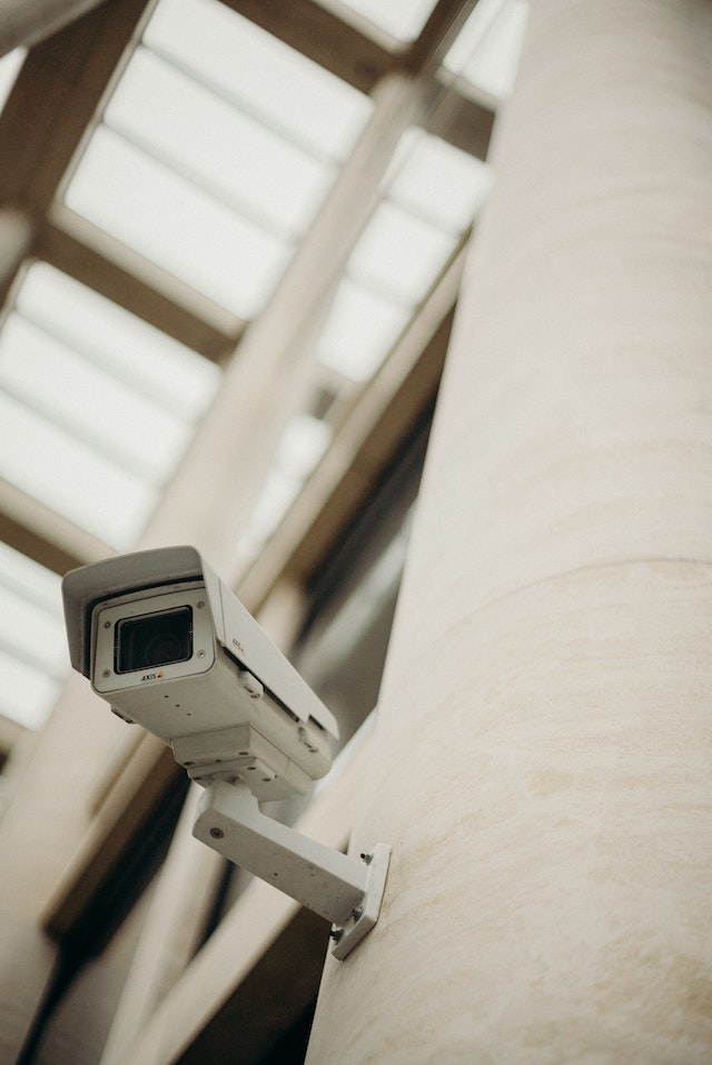 A white security camera on a wall looking down.